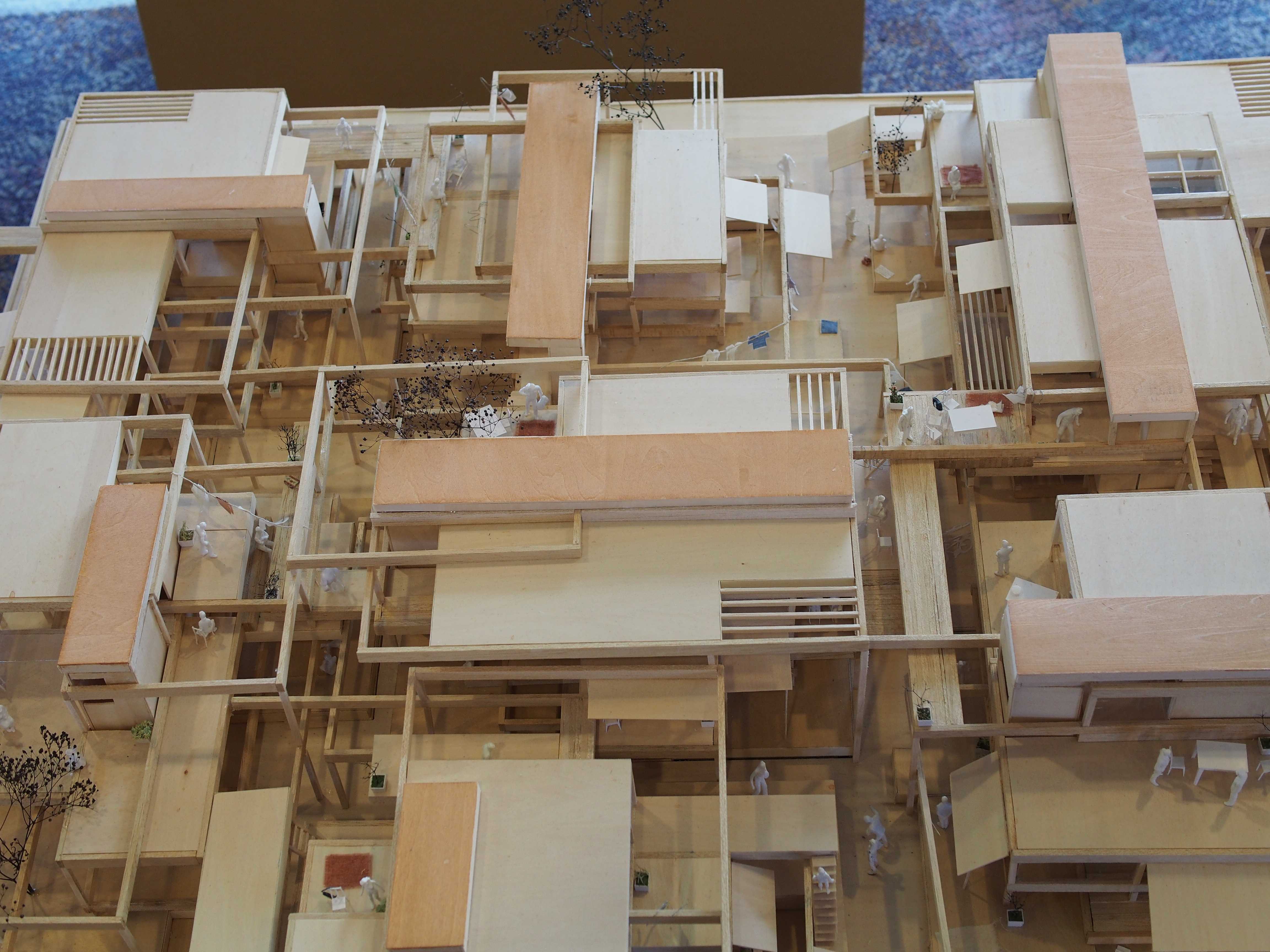 Detailed wooden architectural model of a multi-level complex with small figures, plants, and balconies, in warm tan and wood tones